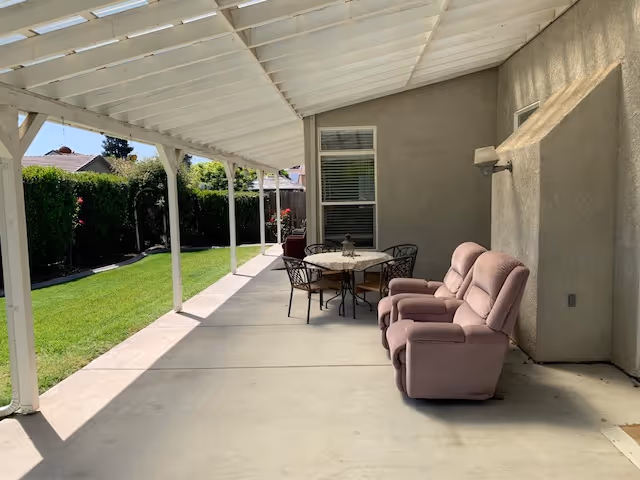 Covered outdoor patio area with two pink cushioned armchairs and a round table with four metal chairs. The patio overlooks a green lawn bordered by hedges and flowers, with a house wall and window in the background.