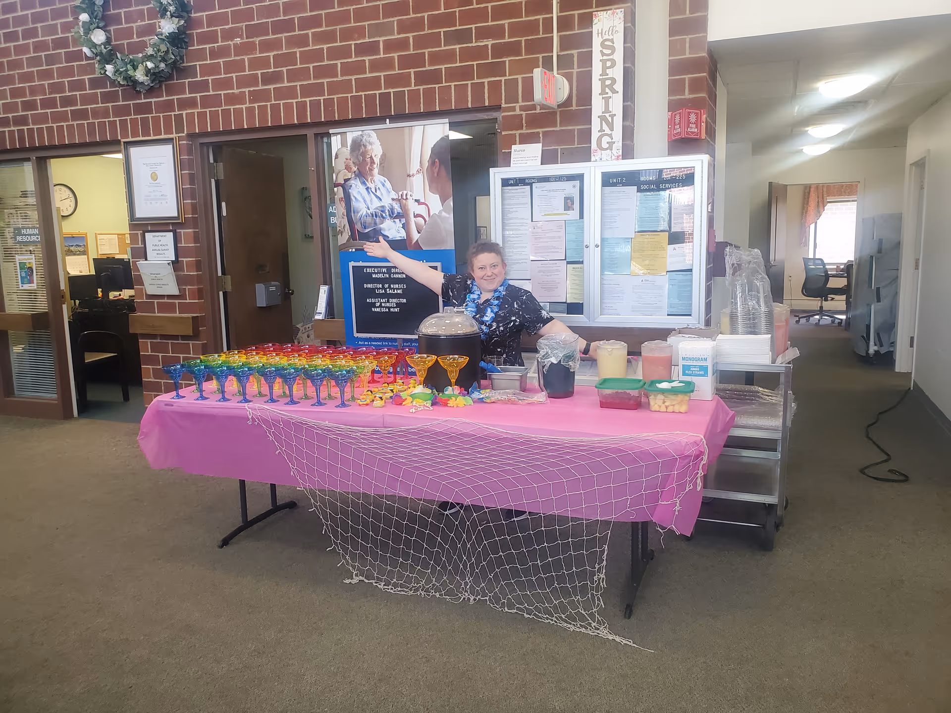A woman stands behind a table covered with a pink tablecloth and decorated with a fishing net. The table holds colorful plastic glasses, drink dispensers, and containers with snacks. The setting appears to be an indoor common area with brick walls, bulletin boards, and office doors in the background. A wreath hangs on the brick wall above the table.