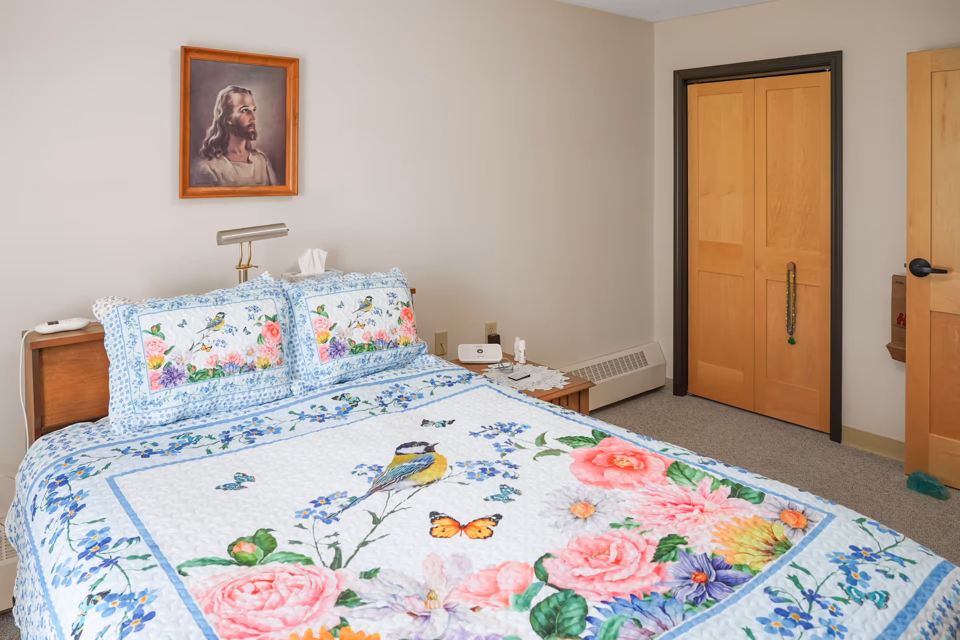 A bedroom with a bed covered in a colorful quilt featuring birds, butterflies, and flowers. Above the bed is a framed portrait of Jesus. There is a wooden nightstand with a lamp, tissues, and a small device. The room has beige walls, a carpeted floor, a wooden closet with sliding doors, and a wooden door with a black handle.