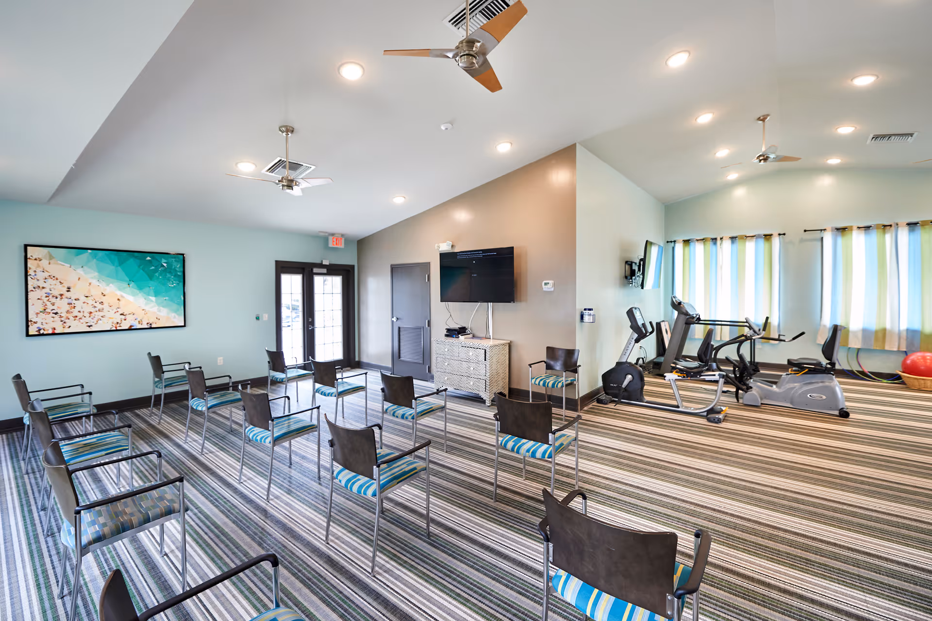 A spacious room with striped carpet flooring featuring rows of chairs with blue and green striped cushions arranged facing a wall-mounted TV. The room has light blue and beige walls, ceiling fans, recessed lighting, and large windows with striped curtains. On one side, there are exercise machines including stationary bikes and elliptical trainers. A large framed picture of a beach scene hangs on one wall near double glass doors.