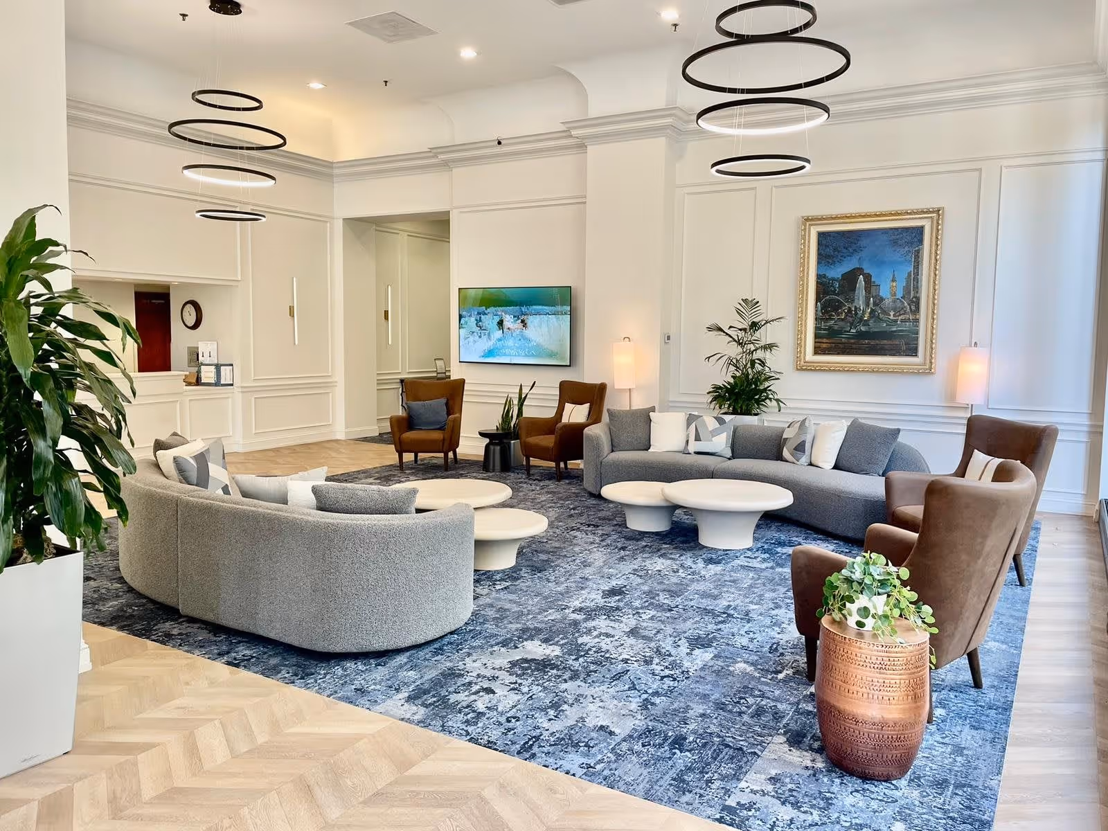 A modern and spacious living room area with a large curved gray sofa, two brown armchairs, and a gray couch arranged around three white round coffee tables on a blue patterned rug. The room features white walls with decorative molding, a large framed painting, two floor lamps, a flat-screen TV mounted on the wall, and several green plants. Circular pendant lights hang from the ceiling, and the floor is a light wood herringbone pattern.