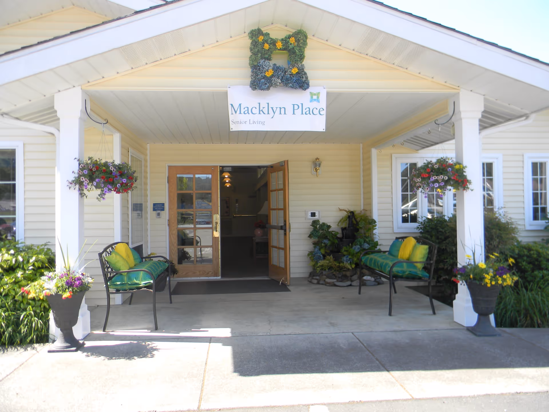 Entrance to Macklyn Place Senior Living with a covered porch featuring two benches with green cushions and yellow pillows, hanging flower baskets, potted plants, and double wooden doors open to the interior.