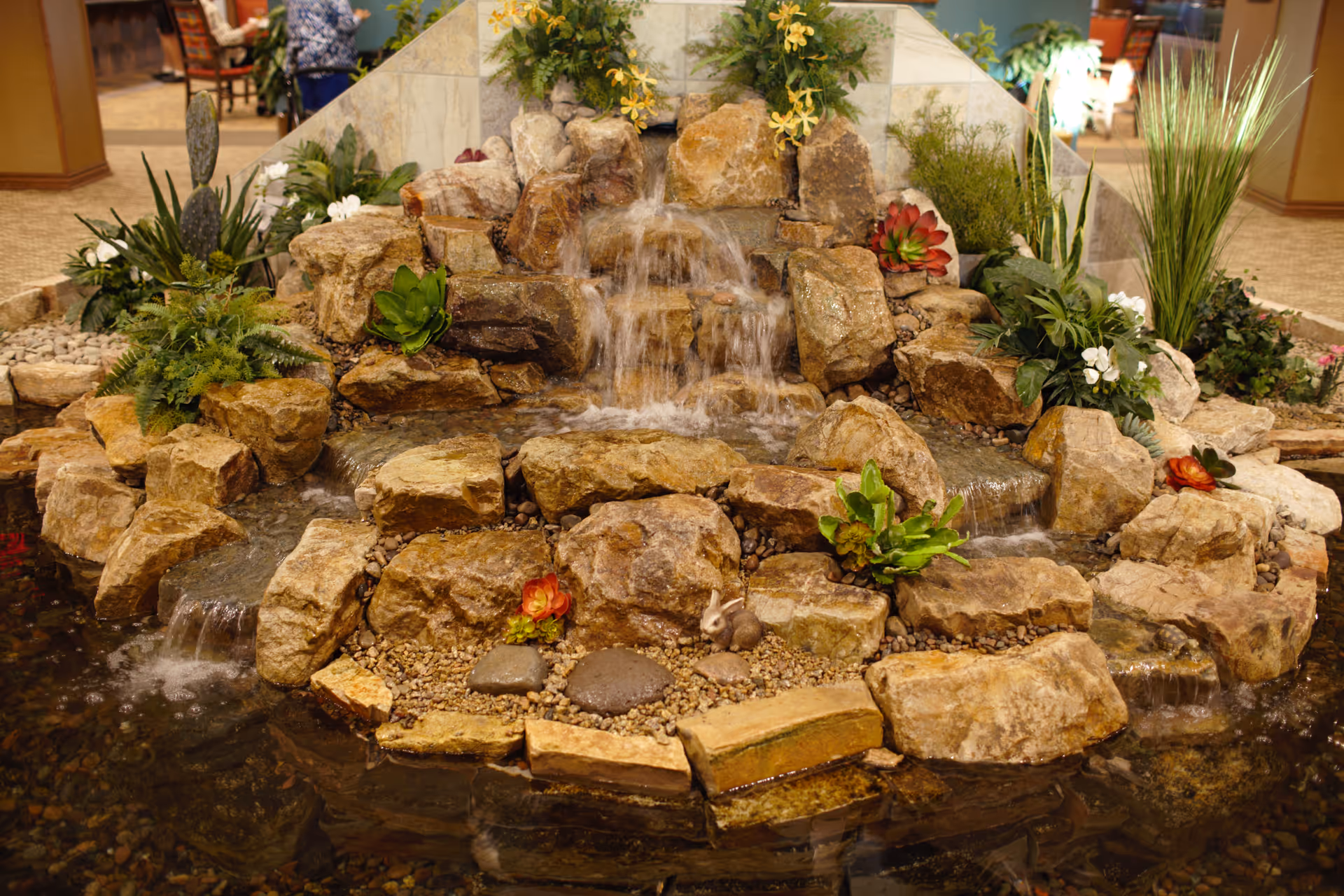 Indoor water feature with cascading waterfall over natural rocks surrounded by green plants and flowers, located inside a building with carpeted floors and seating areas visible in the background.