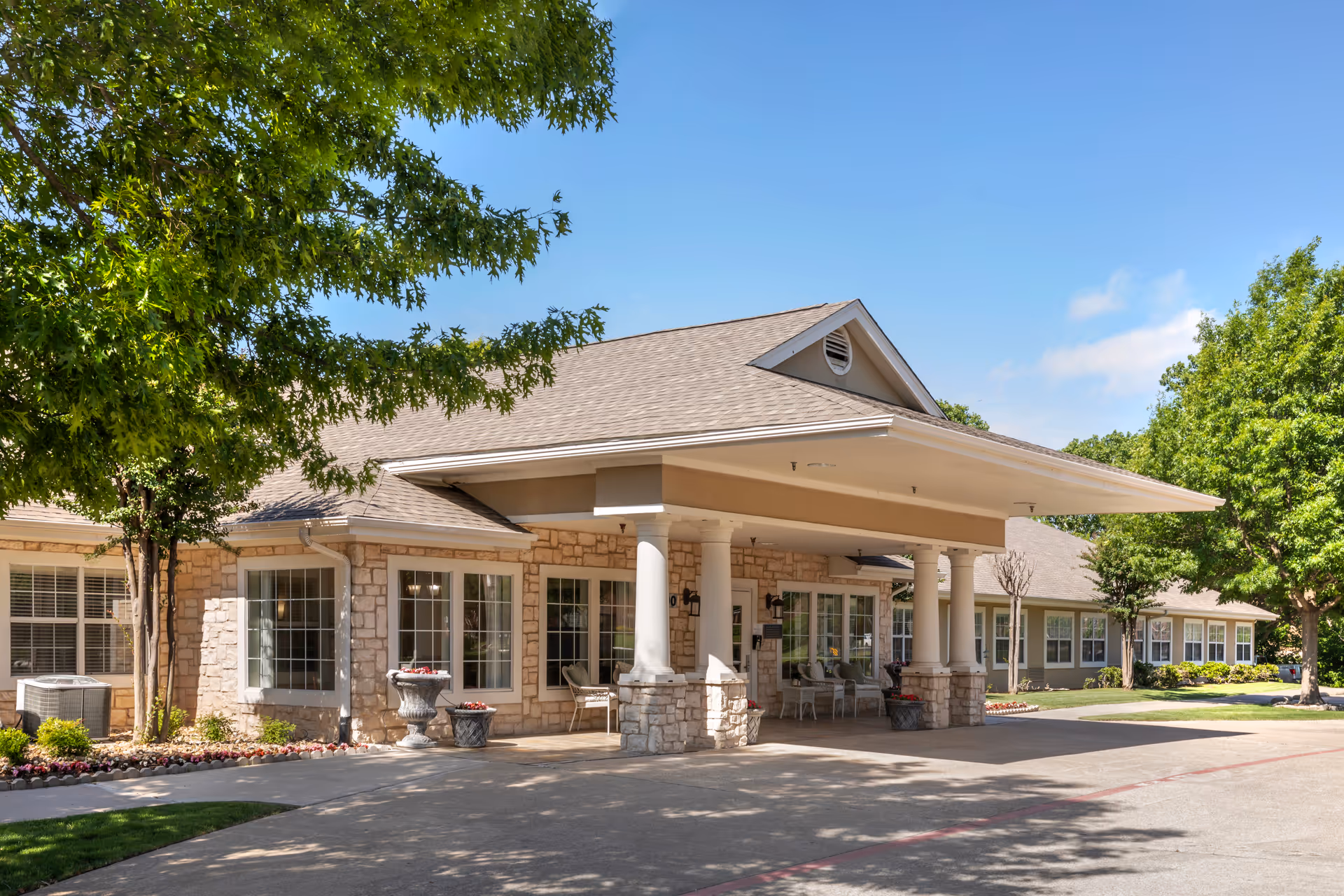 Exterior view of Brookdale Creekside senior living facility showing a single-story building with stone and beige siding, large windows, a covered entrance supported by white columns, and surrounding green trees and landscaping under a clear blue sky.