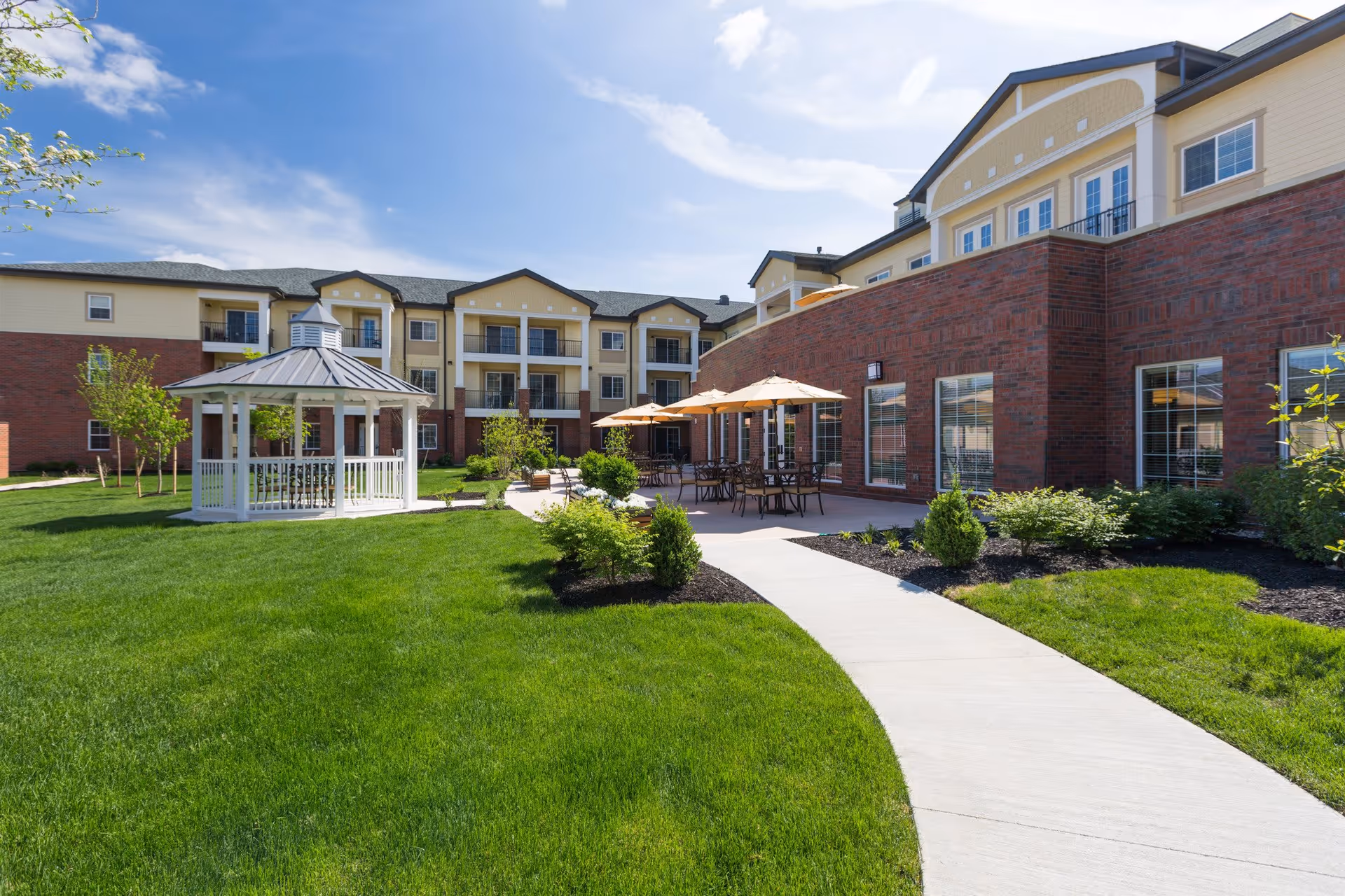 Outdoor garden area of a senior living facility with a white gazebo on the left, a paved walkway, green grass, shrubs, and patio tables with umbrellas next to a multi-story building under a blue sky.