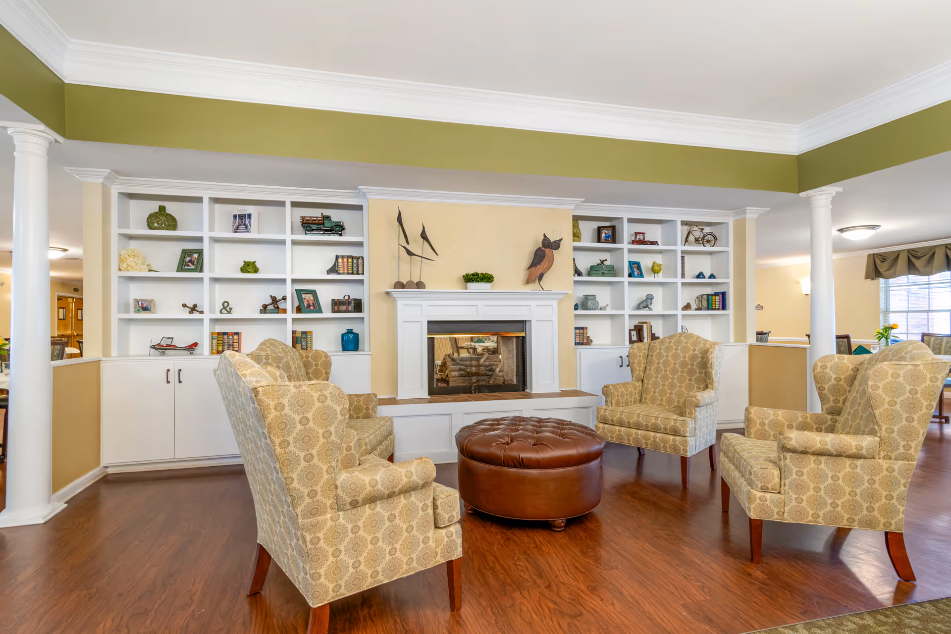 A cozy living room area with four patterned armchairs arranged around a round brown leather ottoman. Behind the seating is a white fireplace with decorative items on the mantel and built-in white shelves filled with books, framed photos, and small decorative objects. The room has wooden flooring, white columns, and a green accent band near the ceiling.