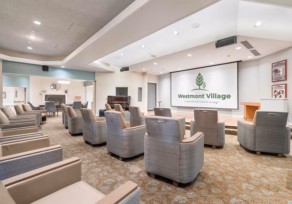 A spacious community room with rows of comfortable armchairs facing a large screen displaying the Westmont Village logo. The room has a patterned carpet, recessed lighting, and a piano near the back. There is an adjoining area with additional seating and a fireplace.