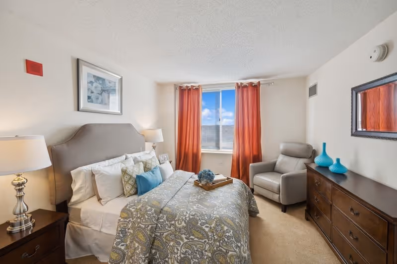 A cozy bedroom with a large bed featuring patterned bedding and multiple pillows. There are two bedside tables with lamps on either side of the bed. A window with red curtains lets in natural light. A gray armchair is positioned next to a wooden dresser with decorative blue vases on top. A framed picture hangs above the bed, and a mirror is mounted on the wall above the dresser.