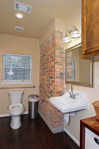 A clean bathroom with a white toilet, a wall-mounted white sink with a silver faucet, a rectangular mirror with a decorative frame above the sink, a brick accent wall, a window with white blinds, a stainless steel trash can, and wooden cabinetry on the right side.