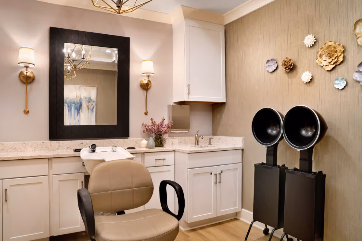 A well-lit salon area with a beige salon chair in front of a white countertop with cabinets. There is a large black-framed mirror on the wall, flanked by two wall sconces with white lampshades. On the right side, there are two black hair dryer chairs with hooded dryers. The wall is decorated with various flower-shaped wall hangings in gold, white, and silver tones.