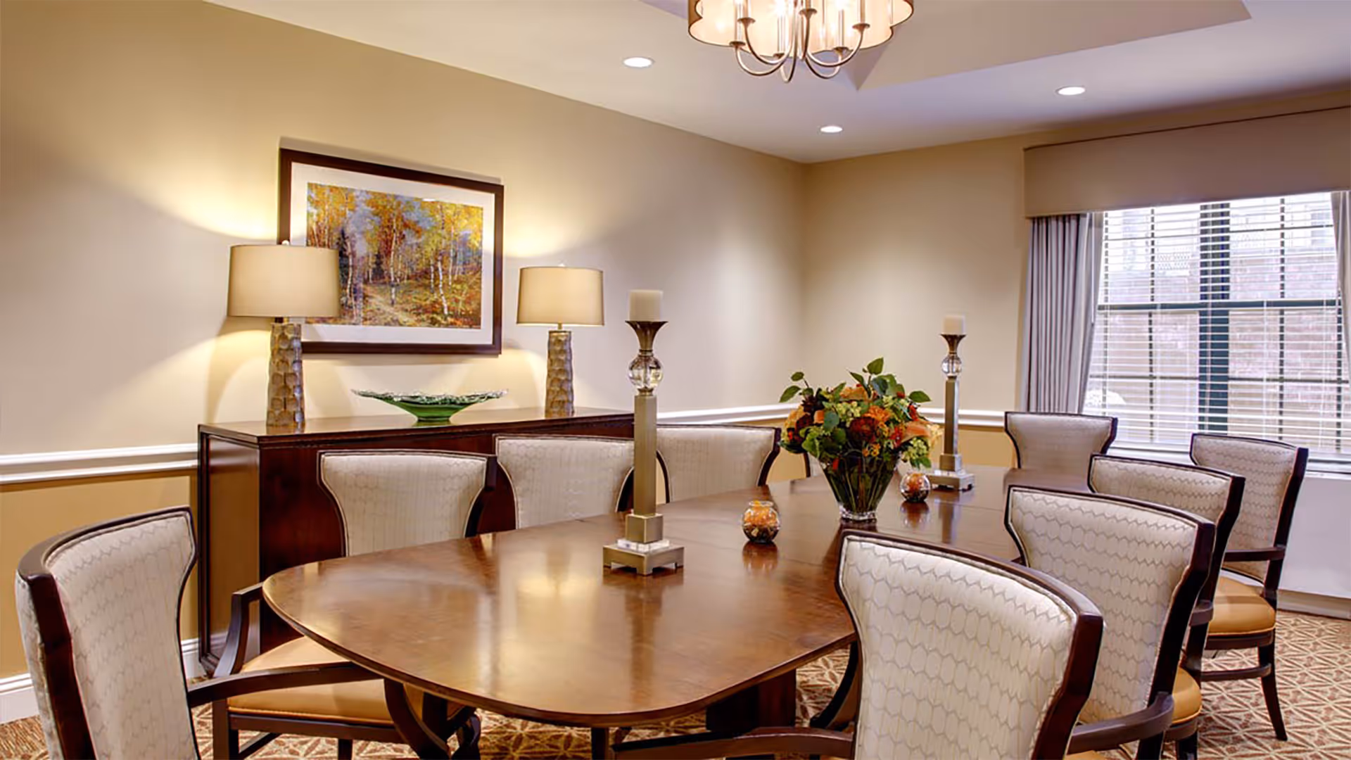 A well-lit dining room with a large wooden table surrounded by eight upholstered chairs. The table is decorated with two tall candle holders, a floral centerpiece, and small decorative items. In the background, there is a sideboard with two lamps and a framed painting of a forest scene on the wall. A window with blinds and curtains allows natural light into the room.