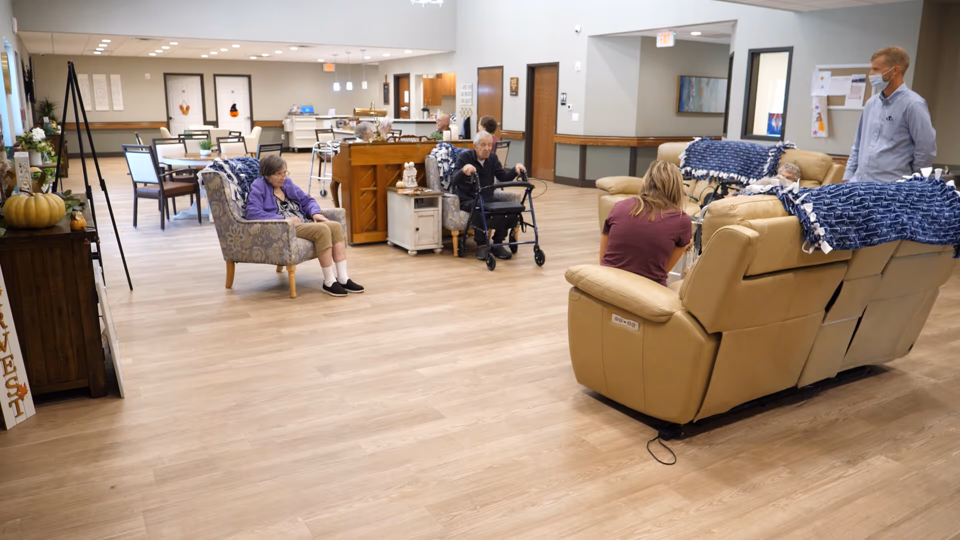 A spacious assisted-living common room with residents seated on chairs and sofas while staff converse nearby.