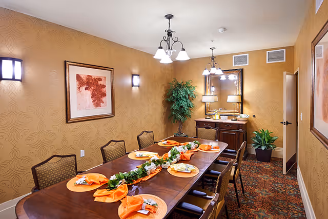 Formal dining room with a long wooden table set with orange napkins and a floral garland, chandeliers, wall art, and a sideboard.