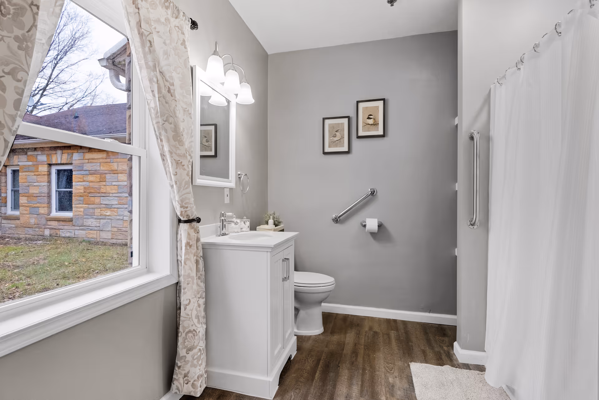 Light gray bathroom with a white vanity and mirror, toilet with grab bars, shower curtain, and a window looking out to a stone building.