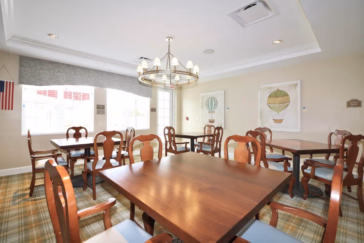 A bright dining room with several wooden tables and chairs arranged neatly. The room features a large window with blinds, two framed pictures of hot air balloons on the wall, a chandelier hanging from the ceiling, and an American flag displayed near the window.