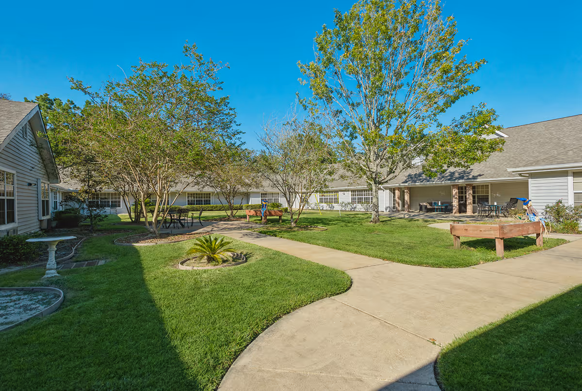 Sunlit courtyard with paved walkways, grassy lawns, trees, benches and patio seating between single-story facility buildings.