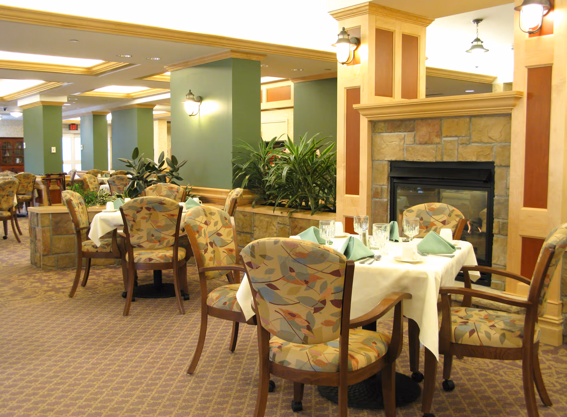 A dining room in a retirement community with tables set with white tablecloths, green napkins, glassware, and silverware. The room features patterned upholstered chairs, a stone fireplace, green pillars, and indoor plants. Warm lighting fixtures are mounted on the walls and ceiling.