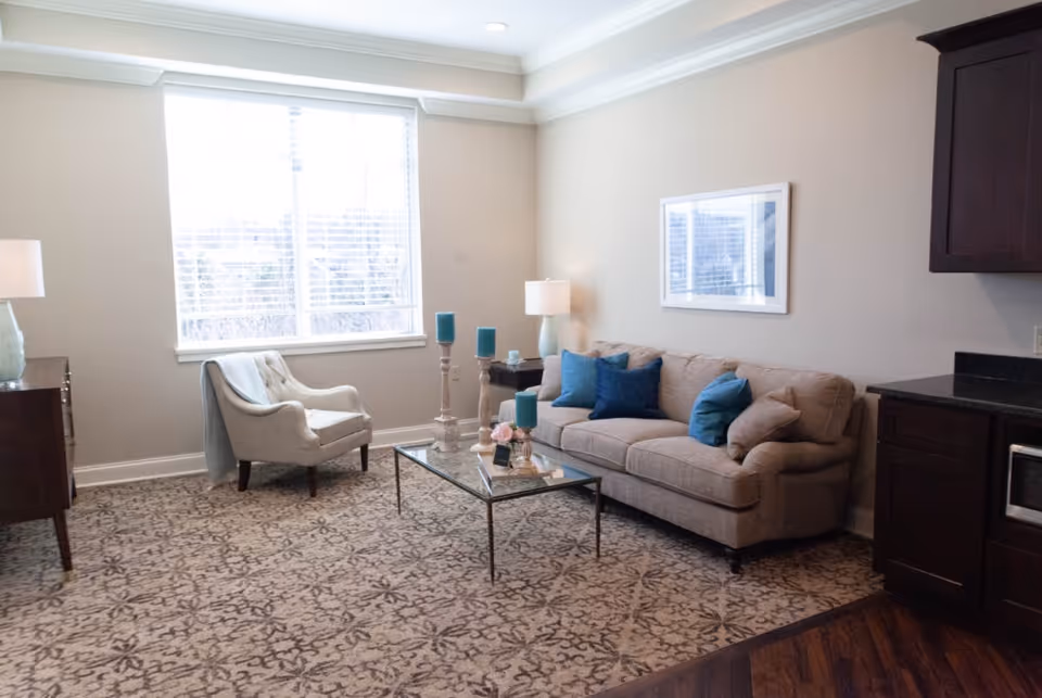 Bright furnished living room with a sofa, armchair, glass coffee table, lamps and a large window.