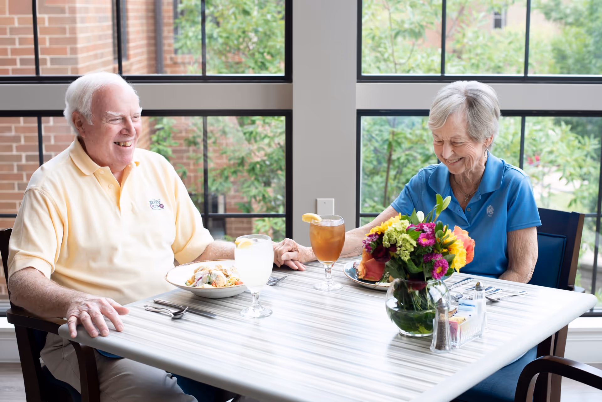 An elderly man and woman sitting at a dining table holding hands and smiling. They have plates of food and glasses of iced tea and lemonade in front of them. A vase with colorful flowers and condiments are also on the table. Large windows with a view of greenery and brick walls are in the background.