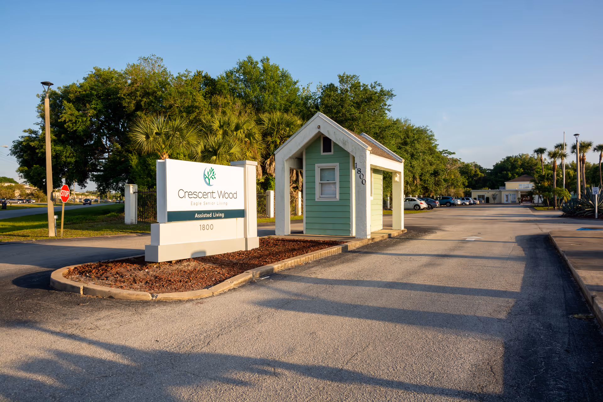 Entrance to Crescent Wood assisted living facility with a white sign displaying the facility name and address 1800, a small green guardhouse, a parking lot with cars, trees, and a clear blue sky.