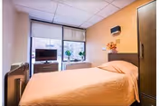A small, cozy bedroom with a single bed covered in a beige bedspread. The room has a large window with a view outside, a flat-screen TV on a stand, and a few decorative plants on the windowsill. The walls are painted in a warm beige tone, and there is a dark wooden wardrobe and headboard. The ceiling has white tiles with recessed lighting.