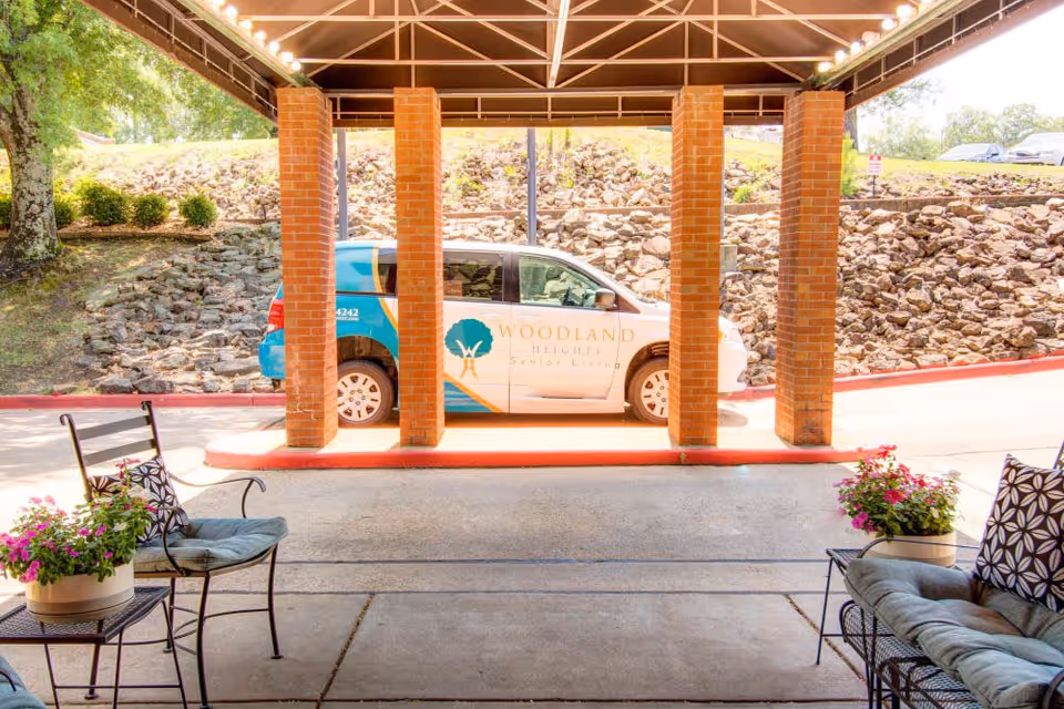Covered entrance area with brick pillars and seating, showing a Woodland Heights Senior Living van parked outside near a rock-lined embankment and greenery.