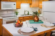 A kitchen with wooden cabinets, white appliances including a microwave, stove, and refrigerator. A kitchen island in the foreground is set with two plates, glasses, and utensils, along with a bowl of fruit and a decorative copper pitcher with greenery.