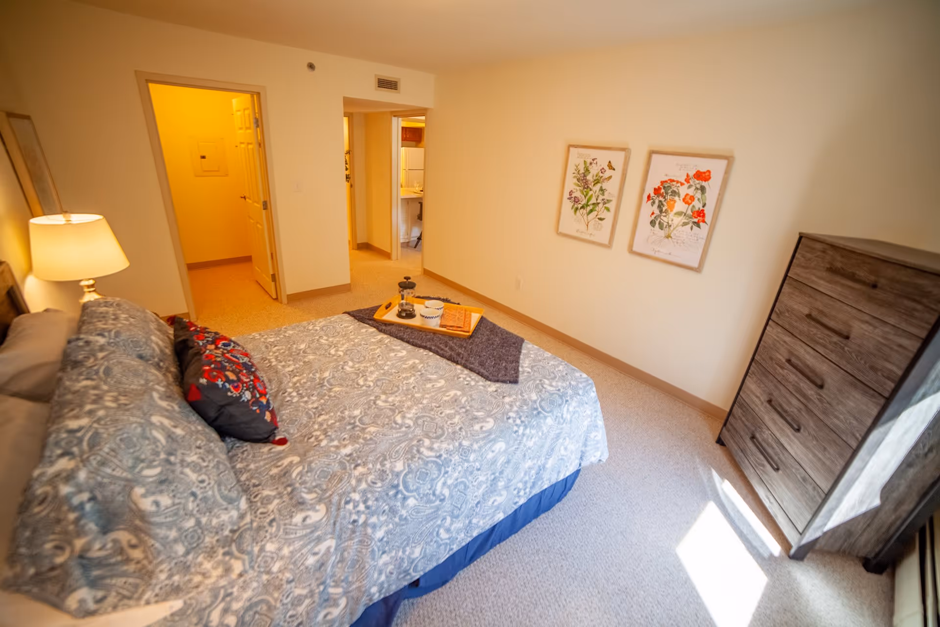 A bedroom with a neatly made bed featuring patterned bedding and decorative pillows. A wooden dresser stands against the wall near a window with sunlight streaming in. Two framed botanical prints hang on the wall. The room has beige walls and carpet, with an open doorway leading to a hallway and another room.