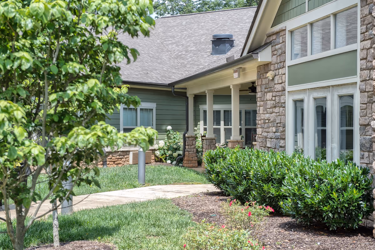 Exterior view of a senior living facility showing a stone and green siding building with large windows, a covered porch with columns, and well-maintained landscaping including bushes, trees, and a walkway.