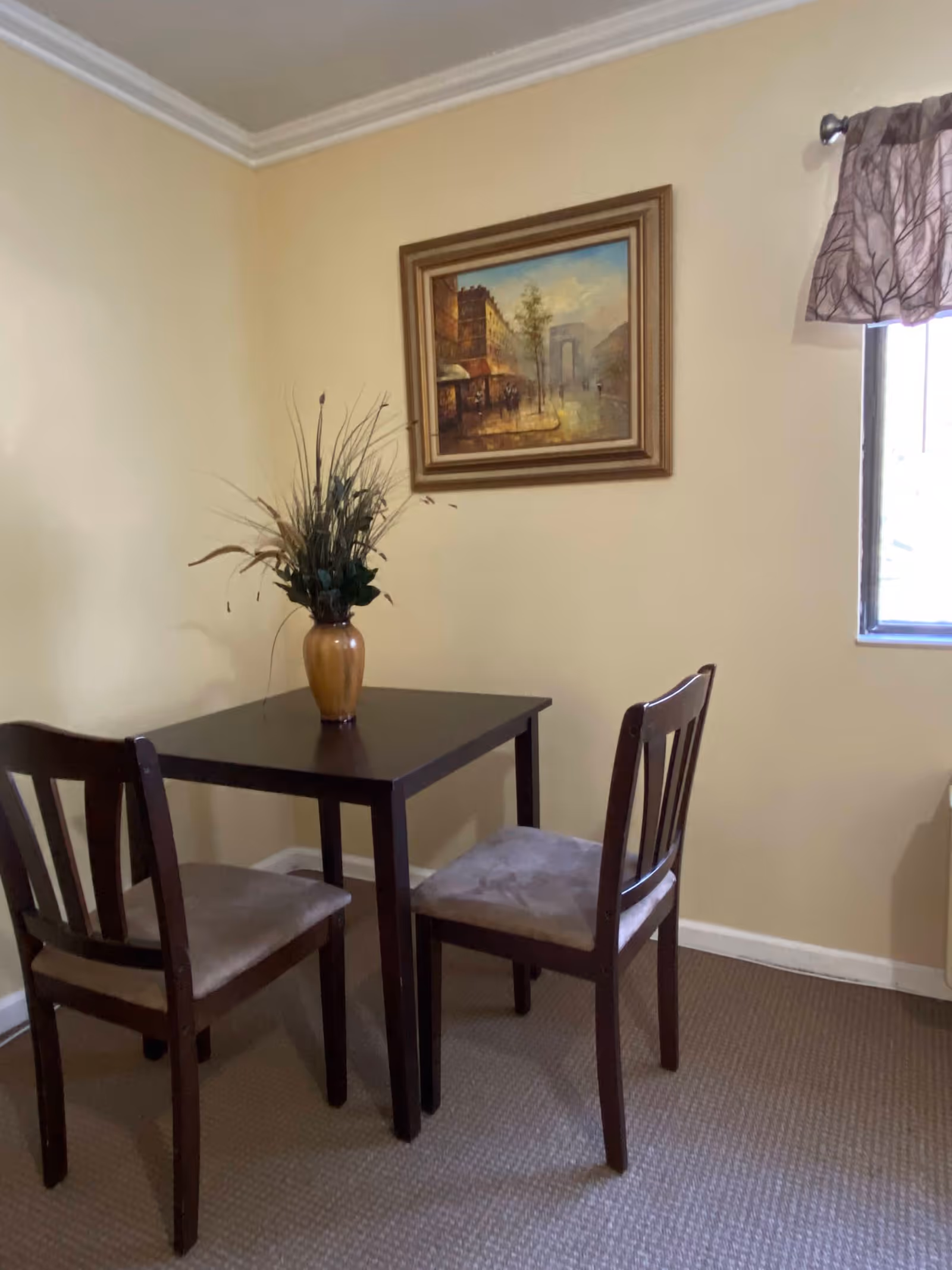 A small dining area with a dark wooden square table and two matching chairs with cushioned seats. A vase with decorative dried plants is placed on the table. The walls are painted light yellow, and there is a framed painting of a street scene hanging above the table. A window with a curtain featuring a tree branch pattern is visible on the right side.