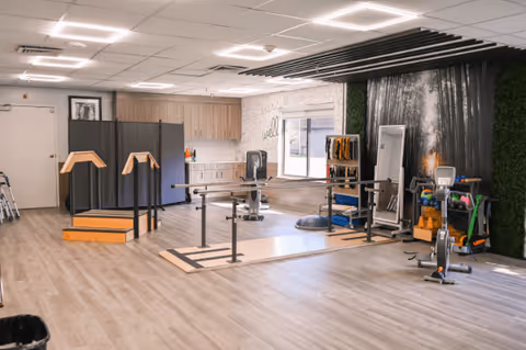A spacious rehabilitation or physical therapy room with parallel bars for walking exercises, a small set of stairs with handrails, exercise equipment including a stationary bike, a large mirror, and shelves with various therapy tools. The room has light wood flooring, a window, and a wall decorated with a waterfall mural and greenery.