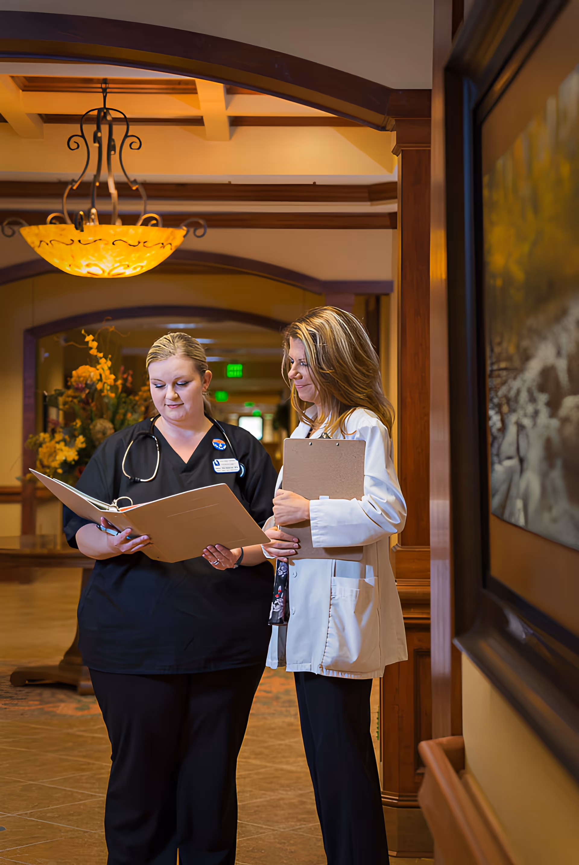 Two healthcare professionals standing in a warmly lit hallway of a senior living facility, reviewing documents together. One is wearing black scrubs with a stethoscope around her neck, and the other is wearing a white lab coat holding a clipboard. The hallway features wooden trim, a decorative chandelier, and floral arrangements in the background.