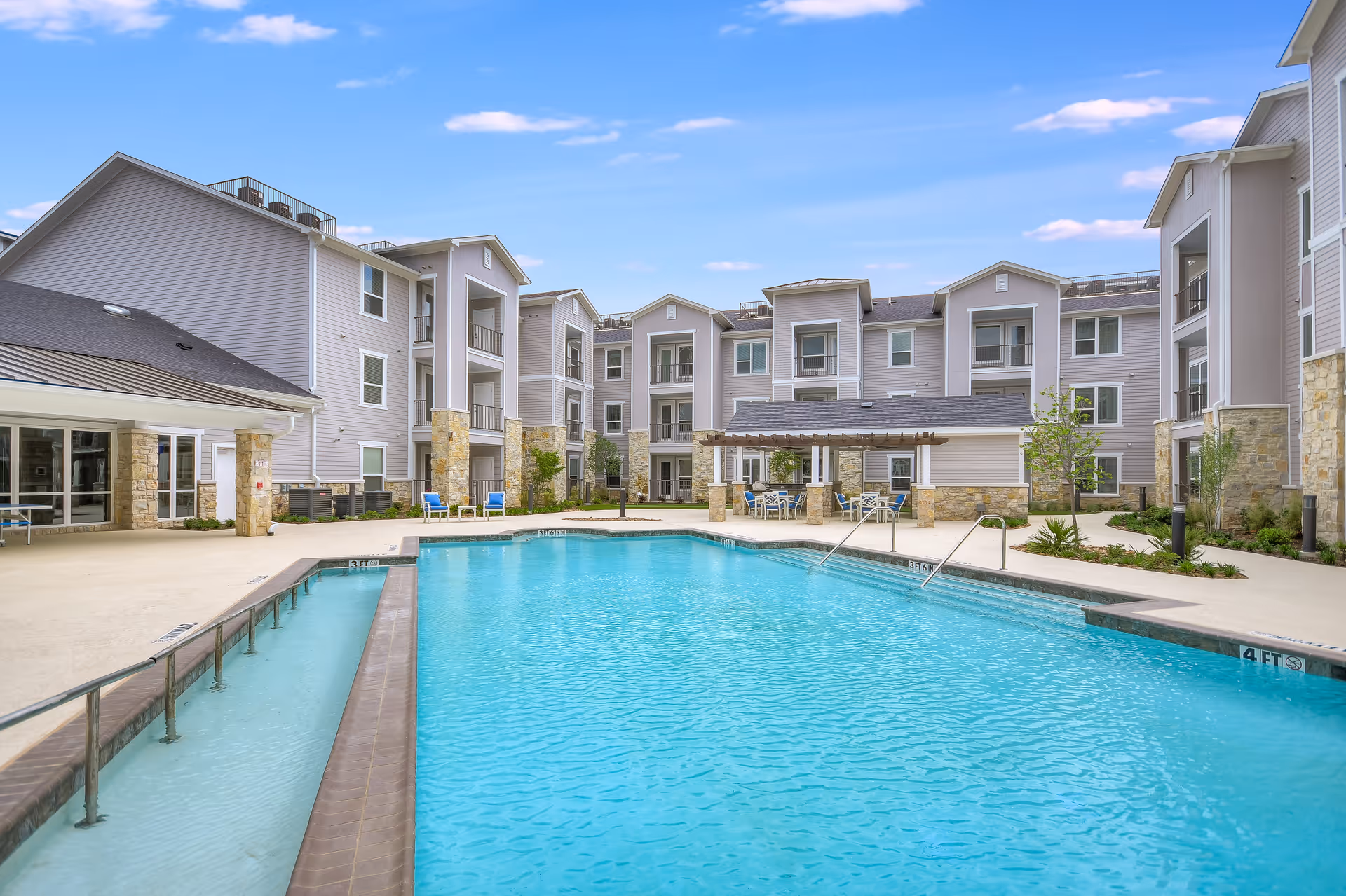 Outdoor swimming pool area at Legacy Oaks of Azle Senior Living with clear blue water, surrounded by a three-story residential building with balconies. There are chairs and tables under a pergola near the pool, and the sky is partly cloudy.