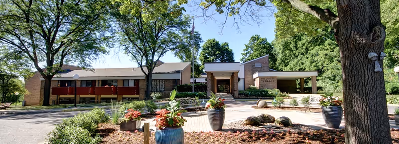 Front exterior of Hillside Terrace Senior Living showing the building entrance, flagpole, trees, and a landscaped courtyard with planters and benches.