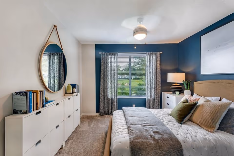 Staged bedroom with a bed, pillows and throw, nightstand and lamp, a dresser topped by a round hanging mirror, and a window with curtains against a blue accent wall.