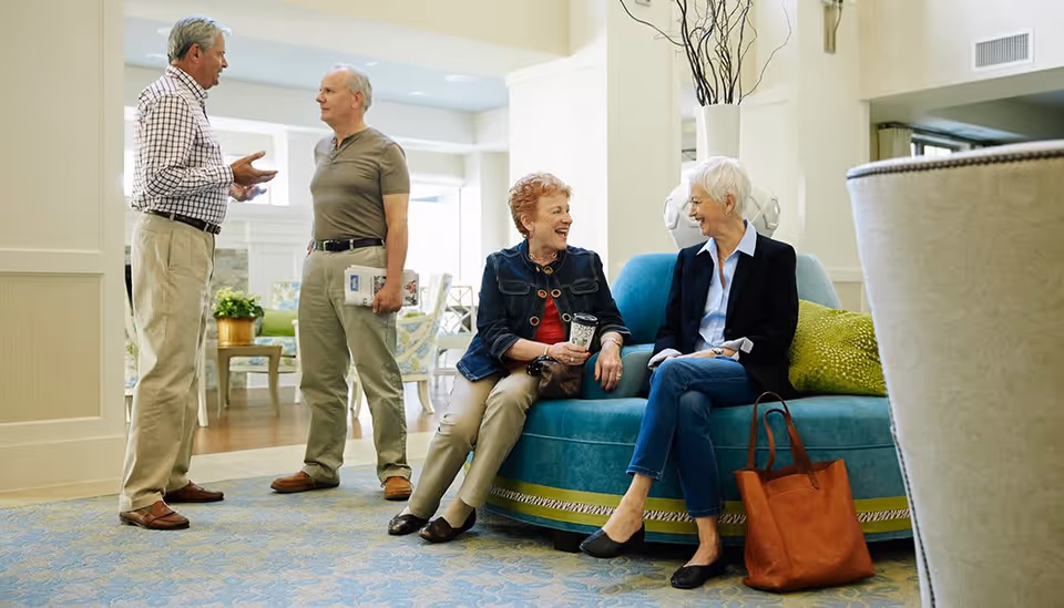 Two elderly women sitting and chatting on a teal couch in a well-lit common area, while two elderly men stand nearby engaged in conversation. The room has light-colored walls, a patterned carpet, and comfortable seating with decorative pillows.