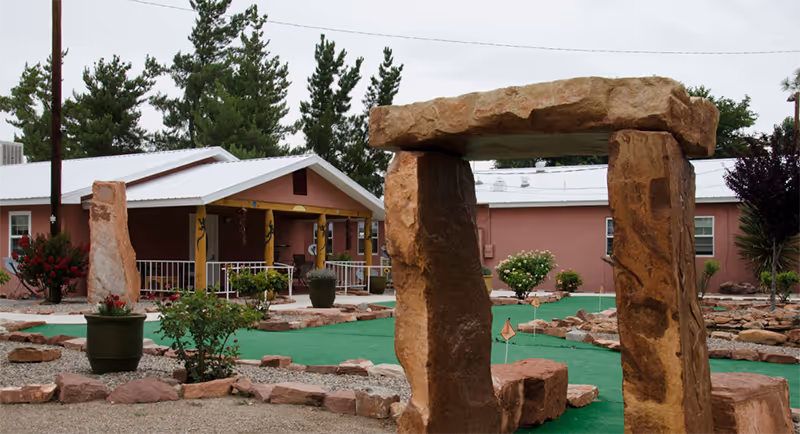 Outdoor area of Casa de Rosa Assisted Living featuring a mini-golf course with green turf and large stone sculptures resembling a stone archway and standing stones. The background shows a single-story building with a covered porch, surrounded by trees and shrubs.