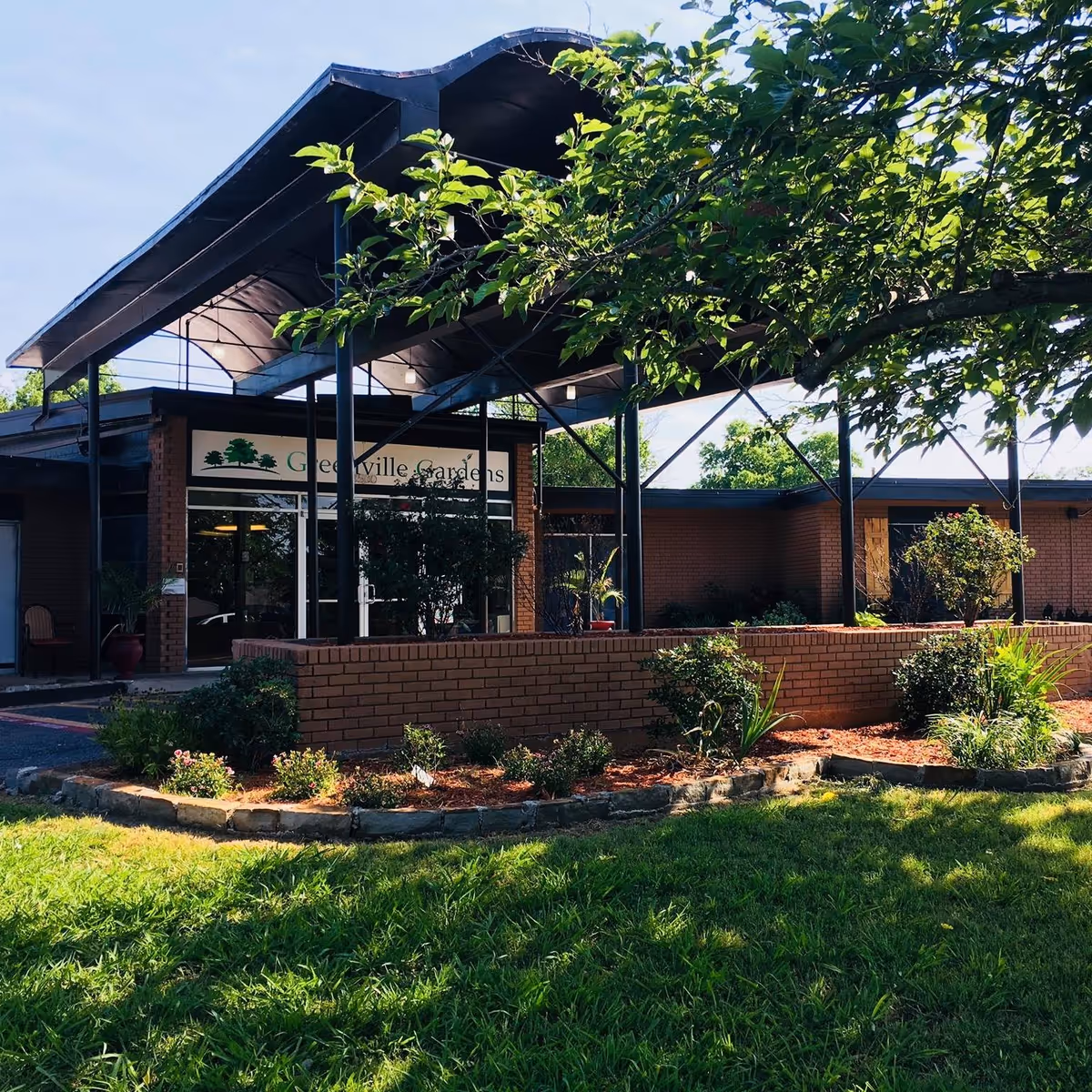 Exterior view of Greenville Gardens facility showing a brick building with a covered entrance, surrounded by green grass, trees, and landscaped plants.