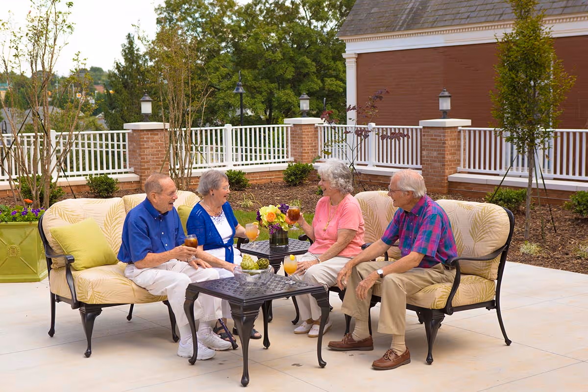 Four older adults sit on outdoor patio sofas at a senior living facility, chatting and holding drinks around a small table.