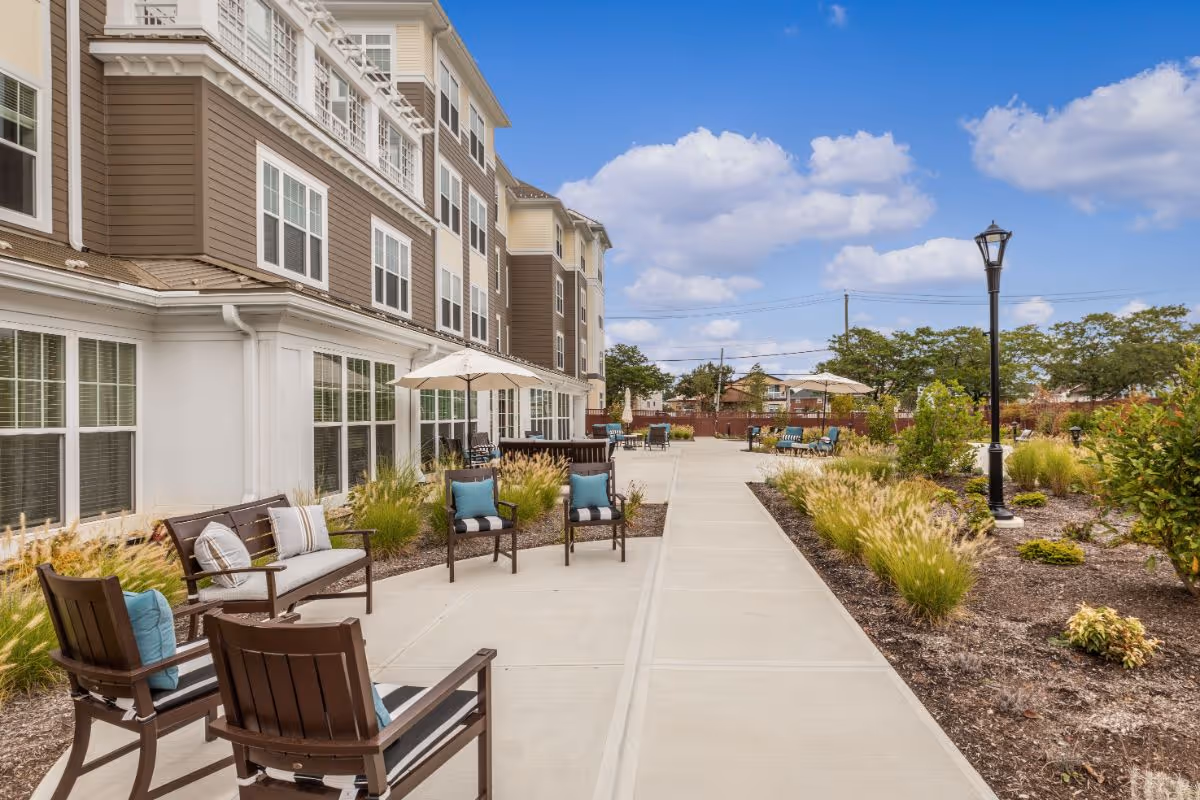 Outdoor patio and walkway with chairs, umbrellas, and landscaping alongside a multi-story senior living building under a blue sky.