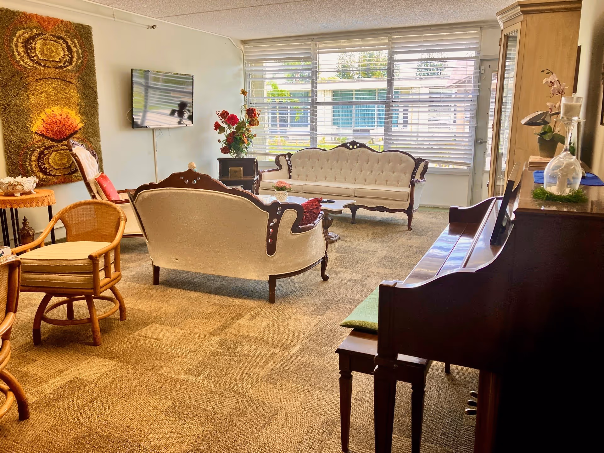 A cozy living room with vintage-style white upholstered sofas and wooden frames, a wooden piano with a bench, a TV mounted on the wall, a large window with horizontal blinds letting in natural light, a decorative wall hanging, and various chairs and floral arrangements.