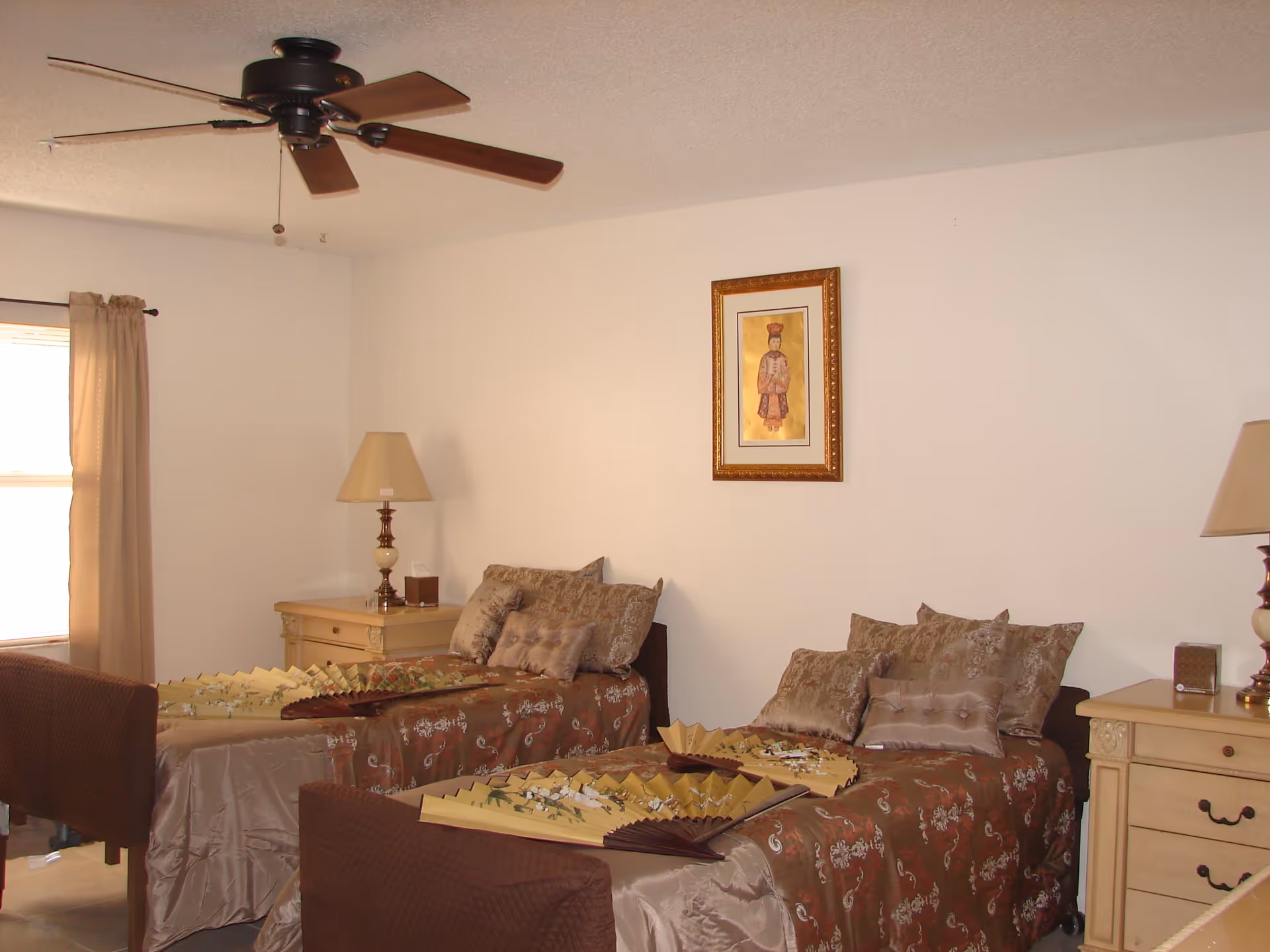 A bedroom with two single beds side by side, each covered with brown and gold patterned bedding and decorative pillows. There are two beige nightstands with matching lamps on either side of the beds. A framed artwork hangs on the white wall above the beds, and a ceiling fan with wooden blades is visible on the ceiling. A window with beige curtains is on the left side of the room.