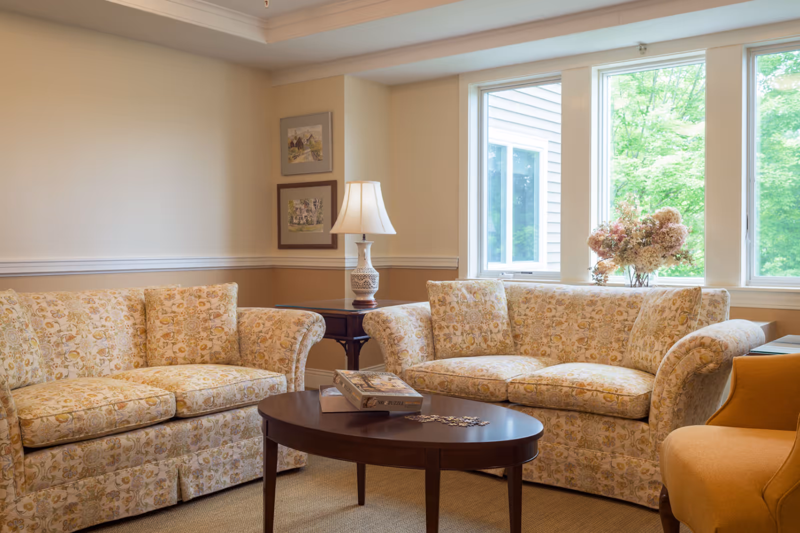 A cozy living room with two floral patterned sofas facing each other, a wooden oval coffee table with a puzzle box and puzzle pieces on it, a side table with a decorative lamp, and large windows letting in natural light with a view of green trees outside. There is also a mustard yellow armchair partially visible on the right.