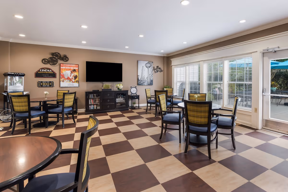 A bright common area with checkered brown and beige flooring, several round tables with chairs, a wall-mounted flat screen TV, decorative wall art, and large windows letting in natural light. There is a popcorn machine on the left and a door leading outside on the right.