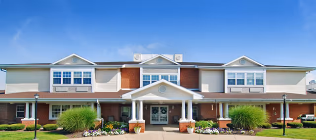 Front exterior view of a two-story senior living facility building with a covered entrance, white columns, brick and beige siding, landscaped flower beds, and green shrubs under a blue sky with some clouds.