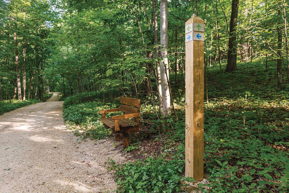 A wooded outdoor trail with a gravel path winding through green trees and foliage. A wooden bench is positioned beside the path, and a wooden post with directional arrows is visible nearby, indicating different trail directions.