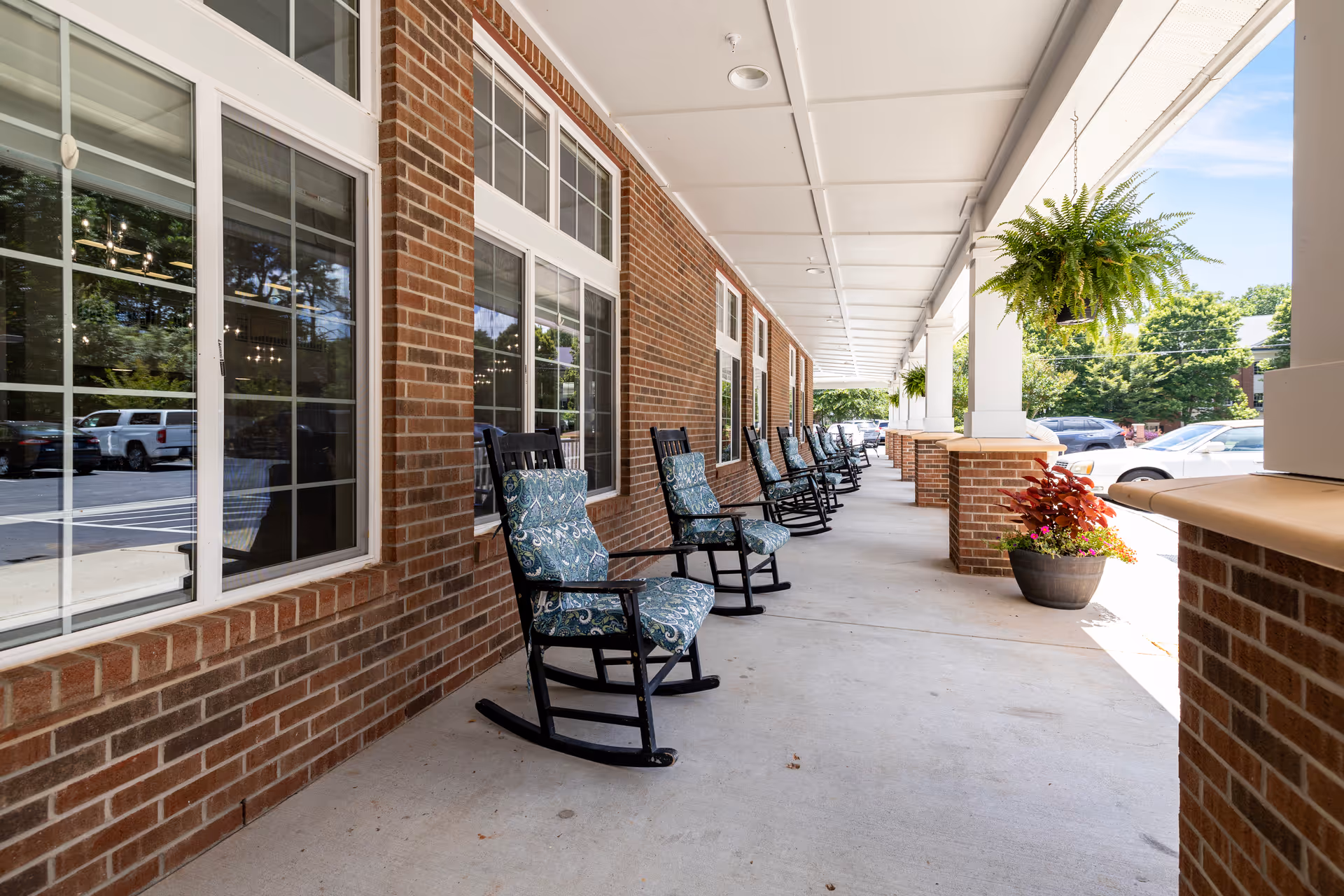 Covered brick-front porch with a row of cushioned rocking chairs, hanging ferns, and potted plants overlooking a parking area.