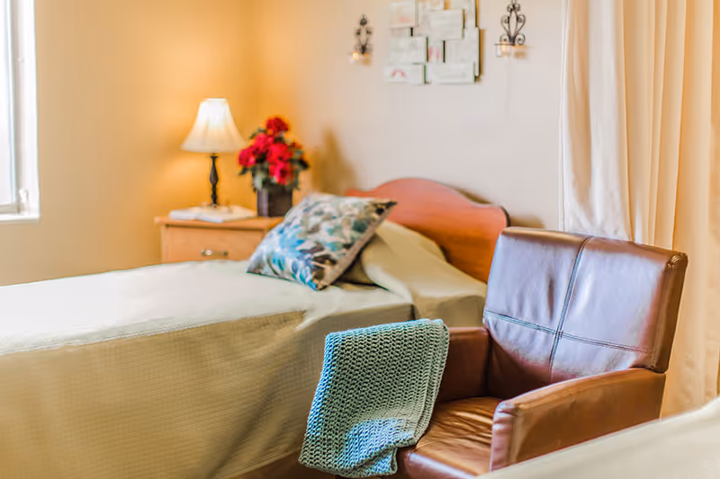 A cozy bedroom with a single bed covered in a light beige bedspread, a decorative pillow, and a wooden headboard. Next to the bed is a wooden nightstand with a lamp and a vase of red flowers. In the foreground, there is a brown leather armchair with a blue knitted blanket draped over the arm. Light beige curtains and a window are visible in the background.