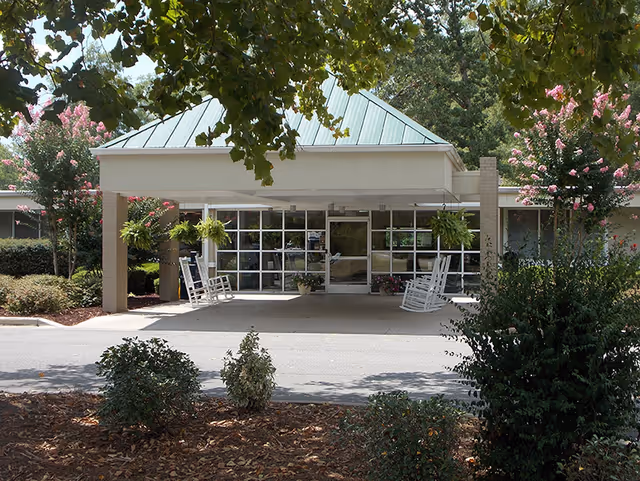 Entrance of McGuffey Healthcare facility with a covered drop-off area, white rocking chairs, hanging plants, and surrounding greenery including trees and bushes.
