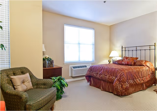 A cozy bedroom with a large window covered by blinds, a bed with a decorative metal headboard and red patterned bedding, a wooden nightstand with a lamp, a green upholstered armchair with a beige pillow, and a potted plant on the floor next to the nightstand.